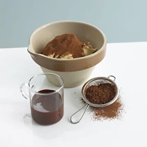 Pudding bowl with butter and cocoa powder next to glass of melted chocolate and sieve of chocolate powder
